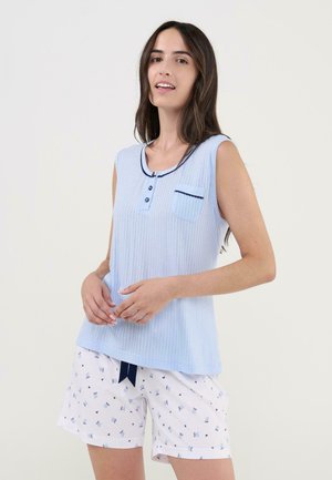 Mujer con cabello largo y oscuro que lleva un top sin mangas azul claro y pantalones cortos blancos con pequeños patrones de lazos azules sobre un fondo liso.