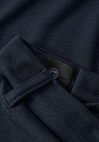 Temno modra tekstura iz šiva, ki vključuje gumb na pasu in zankico za pas. Tkanina ima subtilen tkan vzorec, ki povečuje trdnost.