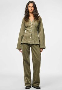 Sieviete ar gariem viļņotiem matiem, tērpusies olīvu zaļā pogājama jakā un atbilstošās platās biksēs, stāvot uz vienkārša gaiša fona.