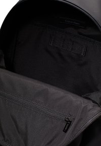 Interno di uno zaino nero con tessuto liscio, dotato di una tasca con zip, un'etichetta e una striscia in velcro per fissare gli oggetti.