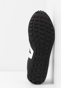 Semelle en caoutchouc noir avec texture antidérapante, tige en tissu sombre avec accents de logo blancs et design épuré.