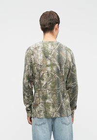 Μακρυμάνικο πουκάμισο με καμουφλάζ σε πράσινο, καφέ και μπεζ χρώμα. Υφασμάτινη υφή με στρογγυλή λαιμόκοψη και εφαρμοστά μανίκια.