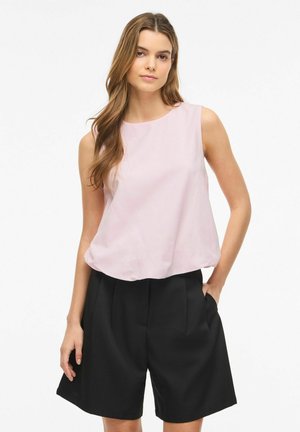 Młoda kobieta z długimi włosami, ubrana w bezrękawnikową bluzkę w bladej różowej barwie i czarne, wysokie szorty z kieszeniami, stojąca na białym tle.