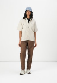 Light beige short-sleeve shirt with two chest pockets, paired with brown trousers and a light blue cap, featuring a logo on the front.