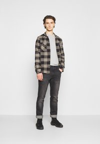 Camisa de franela a cuadros en negro y gris con bolsillos con botones, llevada sobre una camiseta gris clara, combinada con jeans de mezclilla oscura y botas negras.