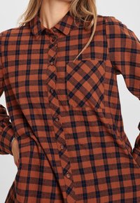 Camicia a quadri marrone e blu navy, con design a bottoni, taschino sul petto e maniche lunghe. Tessuto morbido con motivo a scacchi su tutta la superficie.