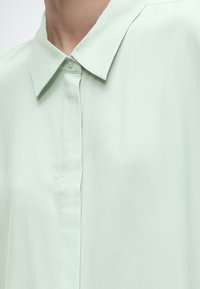 Približen posnetek bledo mintasto zelene srajce z ovratnikom in pokritim gumbnim sprednjim delom, ki jo nosi oseba.