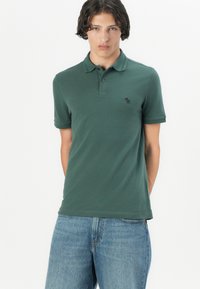 Polo-shirt vert foncé en tissu doux, avec patte de boutonnage à deux boutons et manches courtes. Petit logo sur la poitrine gauche.