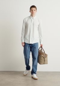 Camisa branca de botões, jeans azuis e sapatilhas brancas. A transportar uma bolsa bege com detalhes castanhos. Fundo plain.