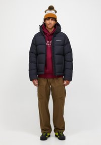 Sort pufferjakke med hætte, maroon hoodie under, brune cargo bukser og fodtøj med gule accenter. Modellen bærer en multikolor hue.