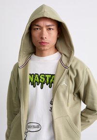 Light green hoodie with a drawstring and red and navy accents. Worn over a white t-shirt featuring bold black and green graphic text.