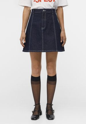 Femme portant une jupe en denim bleu foncé trapèze, des bas noirs transparents montants aux genoux et des chaussures Mary Jane noires, debout sur un fond blanc.