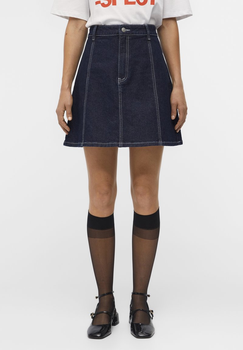 Femme portant une jupe en denim bleu foncé trapèze, des bas noirs transparents montants aux genoux et des chaussures Mary Jane noires, debout sur un fond blanc.