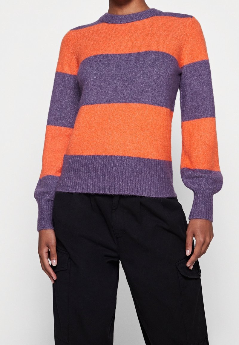 Personne portant un pull à manches longues avec de larges rayures horizontales orange et violettes et un pantalon cargo noir, sur un fond blanc.