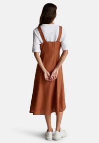 Mujer con un vestido de tirantes color óxido sobre una camiseta blanca y zapatillas blancas, de pie con las manos entrelazadas detrás de la espalda.