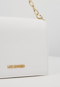 Vit konstläderhandväska med en strukturerad design, guldkedjesnodd och präglad "LOVE MOSCHINO"-logotyp på framsidan. Slät textur.