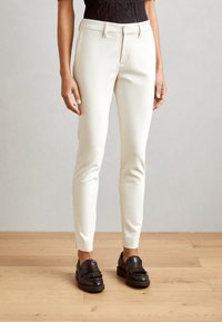 Pantalons blancs ajustés avec une texture lisse, dotés d'une fermeture éclair, de passants de ceinture et de fentes sur les côtés au niveau des chevilles, associés à des mocassins marron.