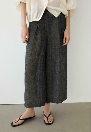 Osoba ubrana w luźne, szerokie spodnie w szarym teksturowanym materiale, kremową bluzkę z podwiniętymi rękawami oraz czarne sandały typu thong, stojąca na szarej podłodze.