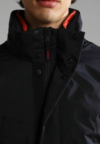 Giacca nera con colletto alto, foderatura interna arancione, zip frontale e bottoni a pressione neri. Presenta una texture liscia e una forma affusolata.