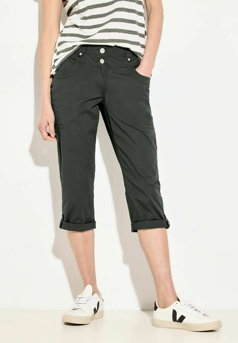 Pantalons courts gris foncé avec ourlets roulés, dotés de poches avant et latérales ainsi que de deux boutons à la taille. Associés à des baskets blanches.