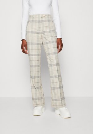 Nainen pukeutuneena beigeihin ruudullisiin räätälöityihin korkeavyötäröisiin housuihin ja valkoisiin paksupohjaisiin tennareihin, seisoo yksinkertaisen taustan edessä.