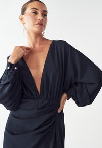 Námornícky šaty s hlbokým V-výstrihom a objemnými dlhými rukávmi. Vyrobené z hladkej tkaniny, s nariaseným detailom v oblasti pásu a perlou gombíkmi ako ozdobou.