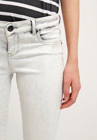 Jeans de mezclilla blanca con un ligero desgastado, diseño de cinco bolsillos y cierre de botón negro, combinados con una blusa a rayas en blanco y negro.
