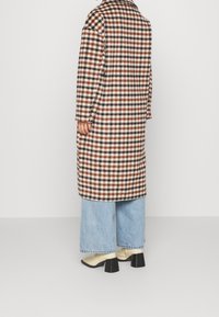 Langer kariert Wollmantel in Braun und Creme mit einem strukturierten Kragen, kombiniert mit weit geschnittenen hellblauen Jeans und beigen Blockabsatzstiefeln.