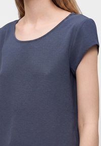 Femme portant un haut gris foncé texturé à manches courtes avec un col rond, montrant la partie supérieure de la poitrine et les épaules.