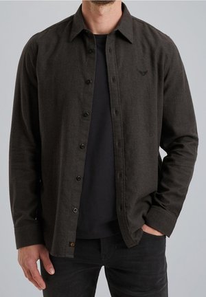 Homme portant une chemise sombre boutonnée sur un t-shirt noir, une main dans la poche, sur un fond clair uni.