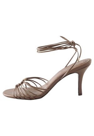 Füße in braunen Sandalen mit Knöchelriemen, sichtbar unter einem plissierten schwarz-beigen gestreiften Rock, stehend auf einem grauen Betonboden.