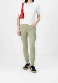 Haut blanc à manches courtes, pantalon cargo vert avec poches latérales et baskets noires avec rayures blanches. Petit sac à main rouge porté à l'épaule.