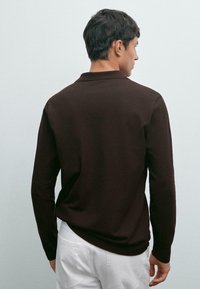 Man facing away wearing a long-sleeve dark brown shirt and white pants against a plain light gray background.