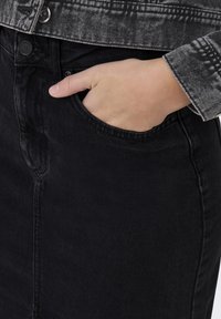 Svart denimkjol med rak skärning, utrustad med en framficka och en delvis synlig grå mönstrad jacka. Ren sömnadsdetalj.