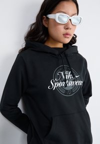 Sweatshirt preto com um logótipo gráfico apresentando "Nike Sportswear" em branco. Capuz anexado, bolso frontal, tecido texturizado. Óculos de sol brancos usados.