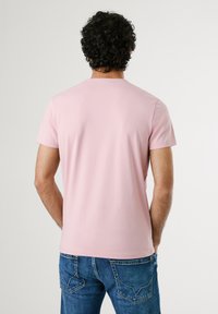 Tricou scurt cu mânecă scurtă, de culoare roz deschis, fabricat dintr-un material moale, având un design simplu cu gât rotund. Este purtat cu blugi albaștri care arată buzunarele din spate.