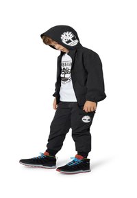 Giacca nera con cappuccio, logo bianco, pantaloni neri con logo, T-shirt bianca e scarpe nere con lacci blu e dettagli rossi.