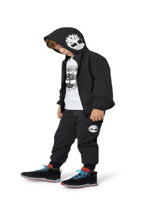 Veste noire avec capuche, logo blanc, pantalon noir avec logo, T-shirt blanc, et chaussures noires avec lacets bleus et accents rouges.