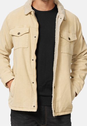 Veste en velours côtelé beige avec doublure en sherpa, poches poitrine à boutons-pression, portée ouverte sur un pull noir, mains dans les poches latérales.