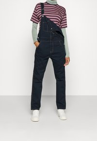 Marineblå denim overalls med justerbare stropper, sidelommer og kontrastsyet. Kombineret med en stribet T-shirt med korte ærmer og hvide sneakers.
