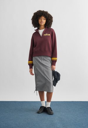 Ung kvinde med krøllet hår iført en vinrød Nike-pullover, grå midi-nederdel, hvide sokker, sorte sko, der holder en sort kasket ved sin side.