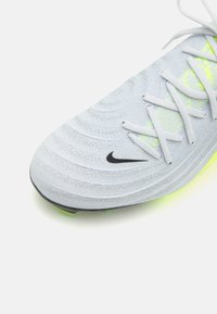 Jasnoszary but sportowy z teksturowaną cholewką, białymi sznurówkami, czarnym logo Nike oraz jaskrawym żółtym akcentem na podeszwie. Okrągły fason palców.