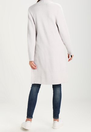 Femme portant un long manteau gris clair, un jean skinny bleu foncé et des baskets blanches, debout de dos face à la caméra sur un fond blanc.