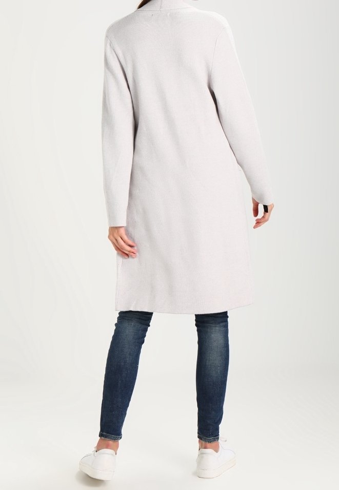 Femme portant un long manteau gris clair, un jean skinny bleu foncé et des baskets blanches, debout de dos face à la caméra sur un fond blanc.