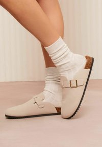 Bežid slip-on kingad ringikujulise ninaga, panuse aktsent ja korgitekstuurne konts, mille alla on pandud valged ribakudumis sokid. Must kummist tald.