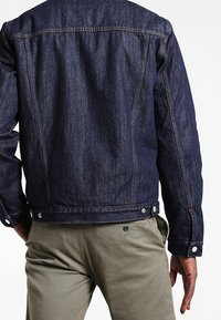 Denim jacket in dark indigo, features visible stitching, snap buttons, and a structured design. Worn over olive green pants.