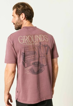 Mann mit Bart, der ein mauvefarbenes T-Shirt trägt, zeigt eine Kaffeekanne, eine Kaffeetasse, einen Bücherstapel und Text über Abenteuer und ein lokales Café.