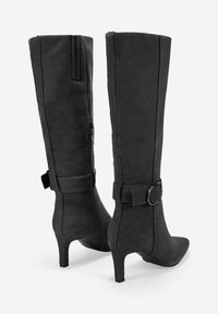 Botas de ante negras hasta la rodilla con puntas puntiagudas, tacones kitten, cremalleras laterales y correas decorativas con hebillas alrededor de los tobillos.