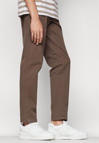 Brown cotton trousers with a straight fit, rolled cuffs, and side pockets, paired with white sneakers. Striped top partially visible.