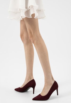 Beine in bordeauxroten Wildleder-Pumps mit spitz zulaufender Spitze, kombiniert mit einem weißen, fließenden Rock vor weißem Hintergrund.